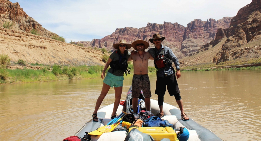 three people stand on a raft and pose for a photo. the raft is floating on water in a canyon.